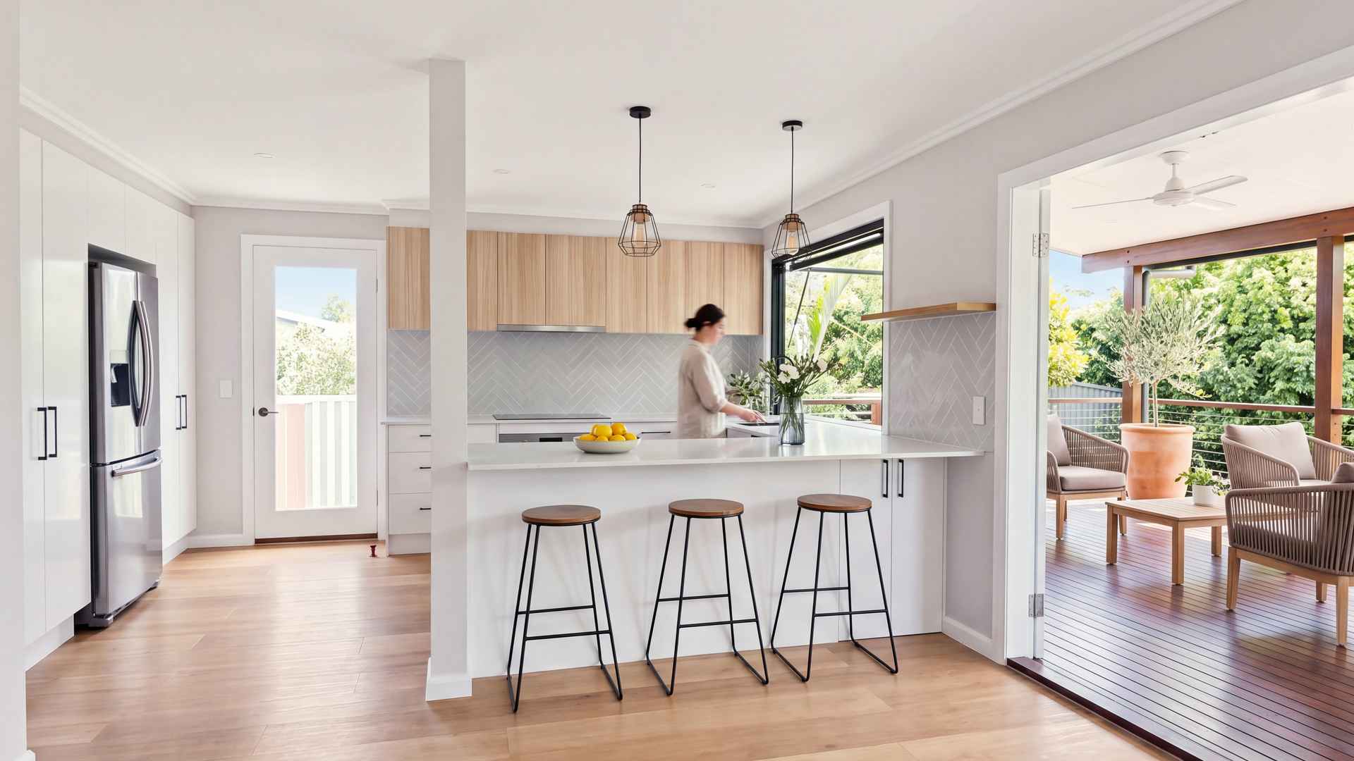 Open-plan kitchen with an island and three stools, opening onto the covered deck in soft daylight
