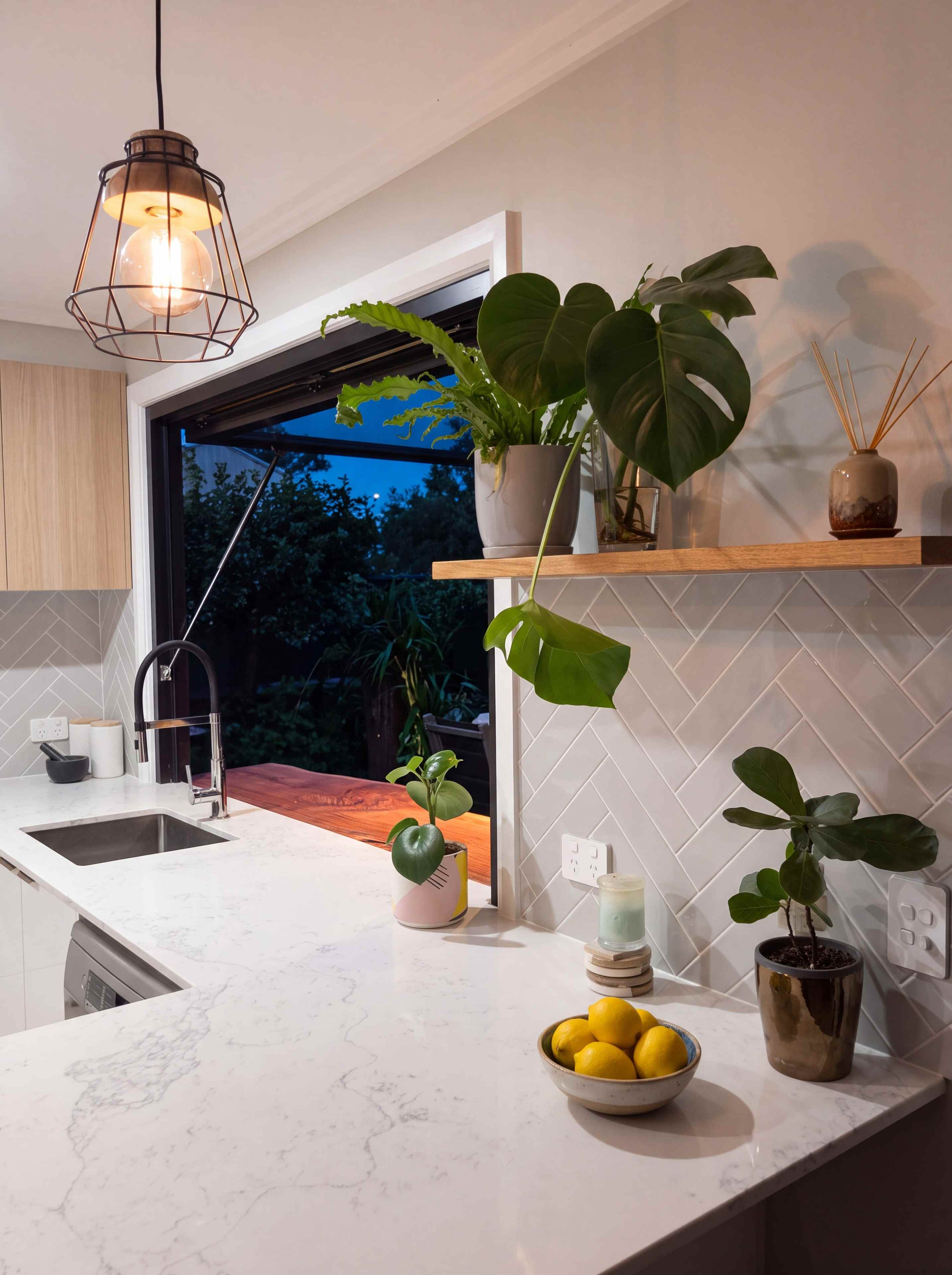 The kitchen at dusk, with a dark bronze cage pendant glowing over the stone benchtop, the servery window open to the evening sky and plants on a floating oak shelf
