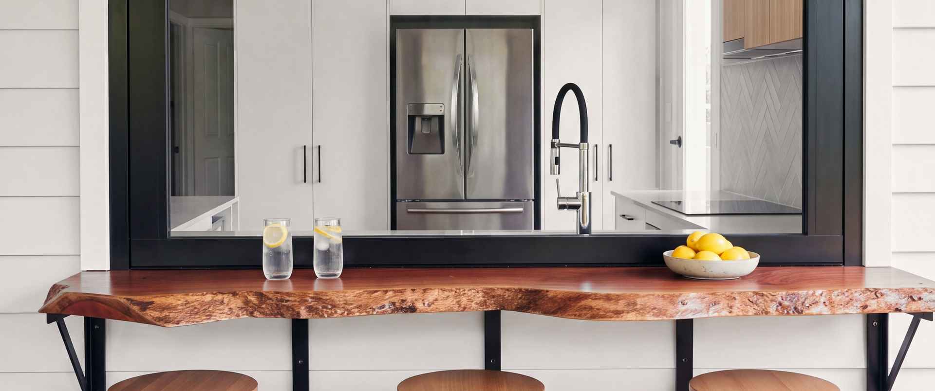 A live-edge red timber bar under the open servery window of the Everton Hills kitchen, with three timber stools on the deck