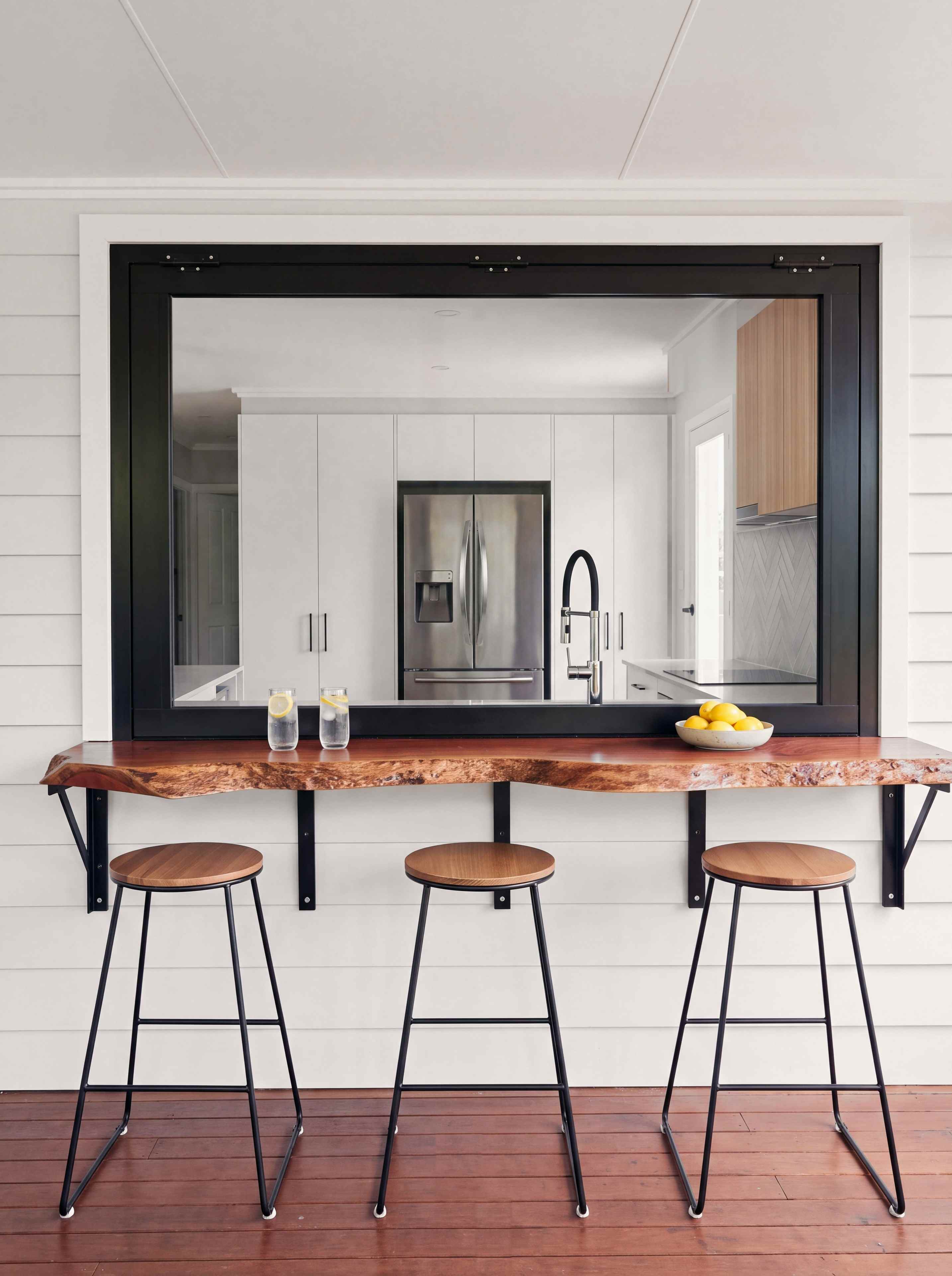 The servery window from the covered deck, with a live-edge red timber bar, three stools and white weatherboard walls