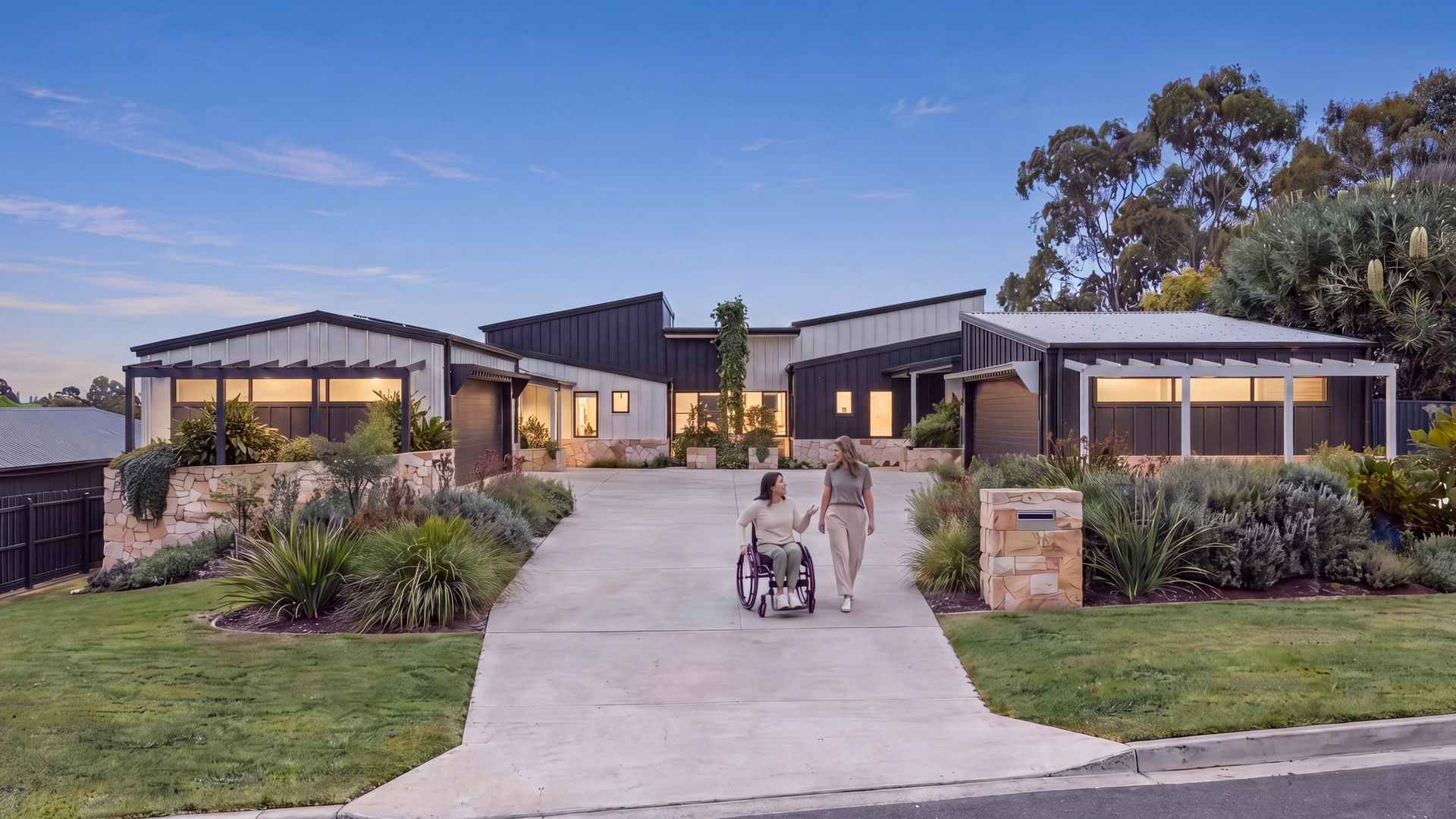 Single-level SDA duplex at dusk with warm lights on, and a resident using a wheelchair with a friend on the level driveway