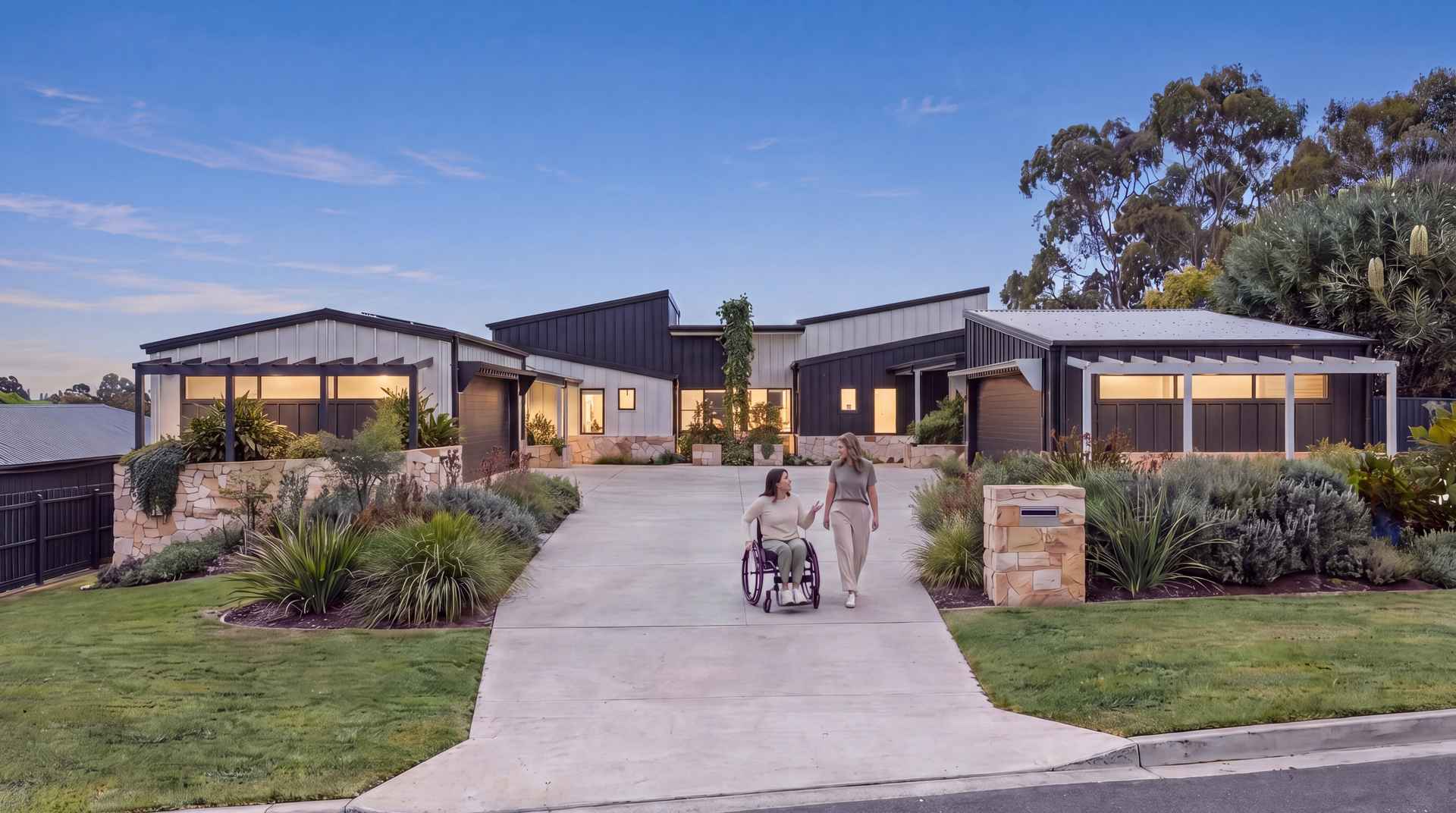 Single-level duplex at dusk with warm lights on, and a woman using a wheelchair with a friend on the level driveway