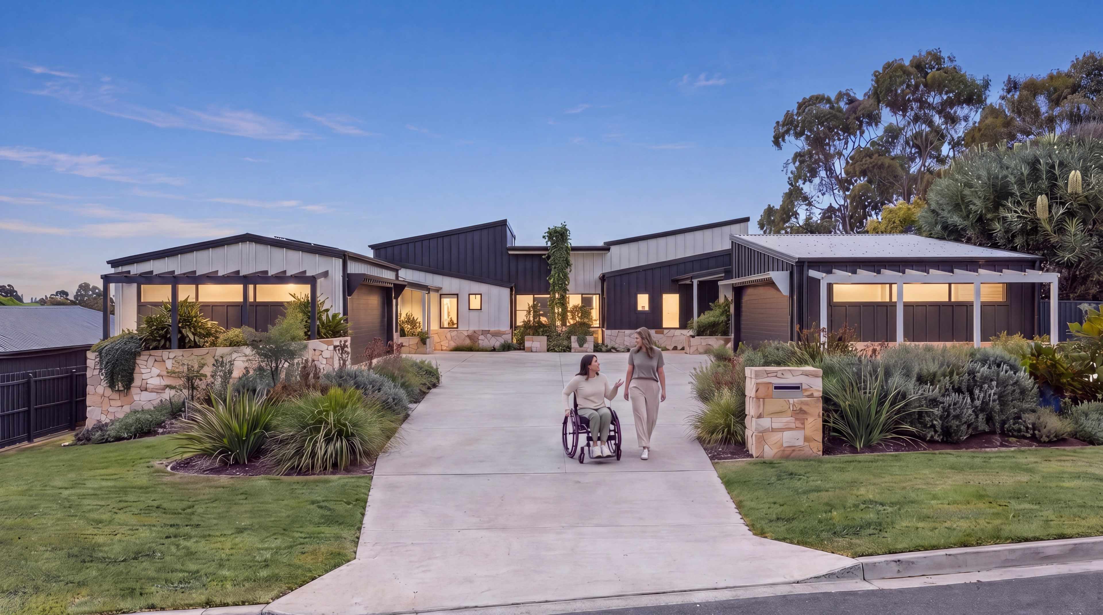 Single-level SDA duplex at dusk with warm lights on, and a resident using a wheelchair with a friend on the level driveway