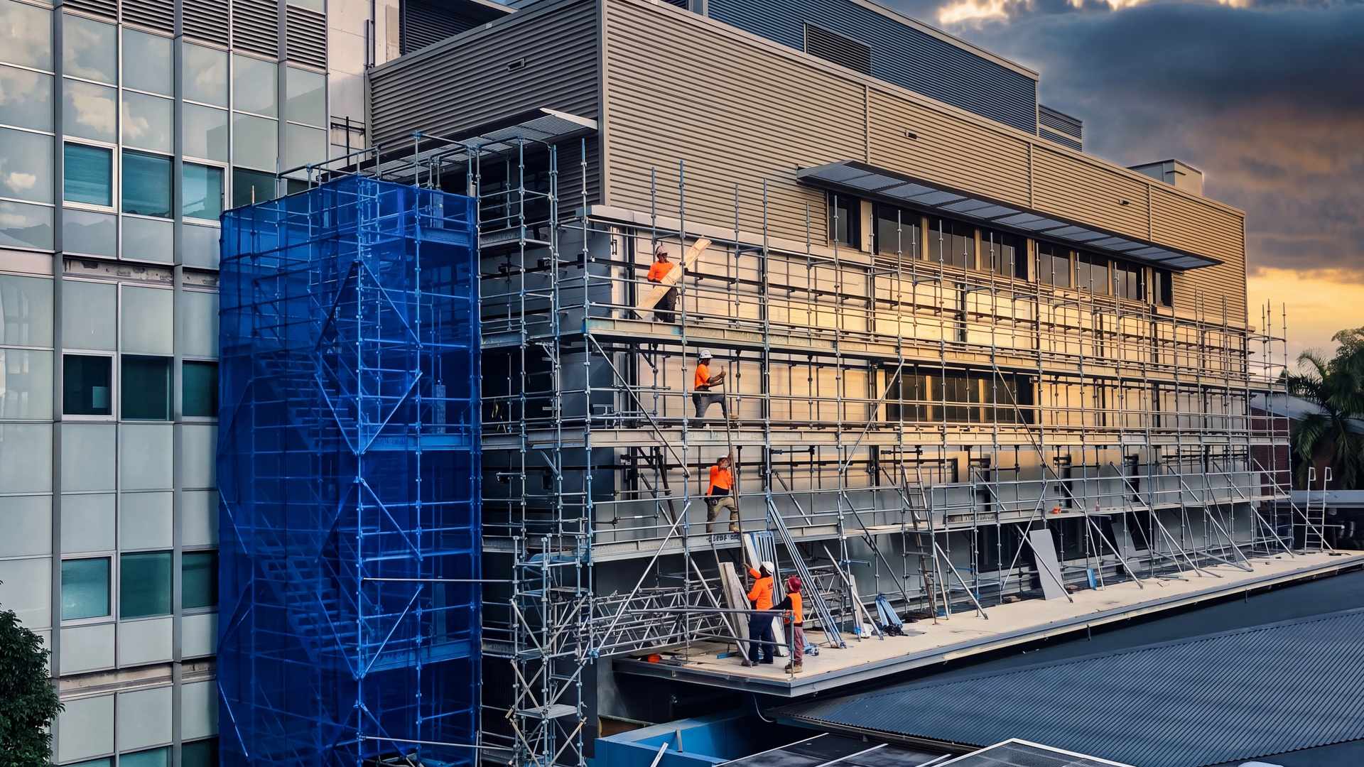 A crew on scaffold passes new facade panels up the side of an operating hospital at golden hour