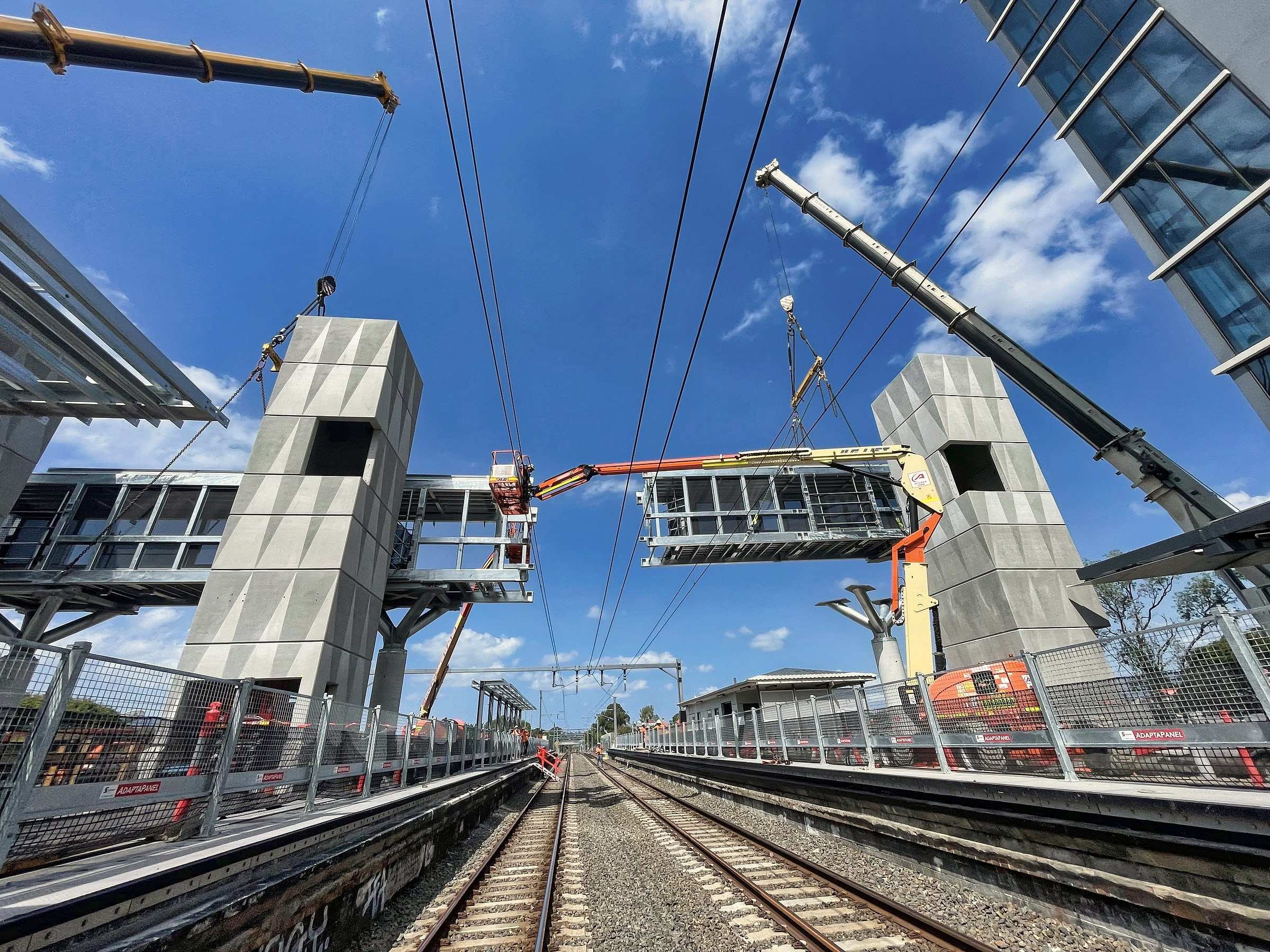 A crane lifting a footbridge section between two station lift towers above the rail line