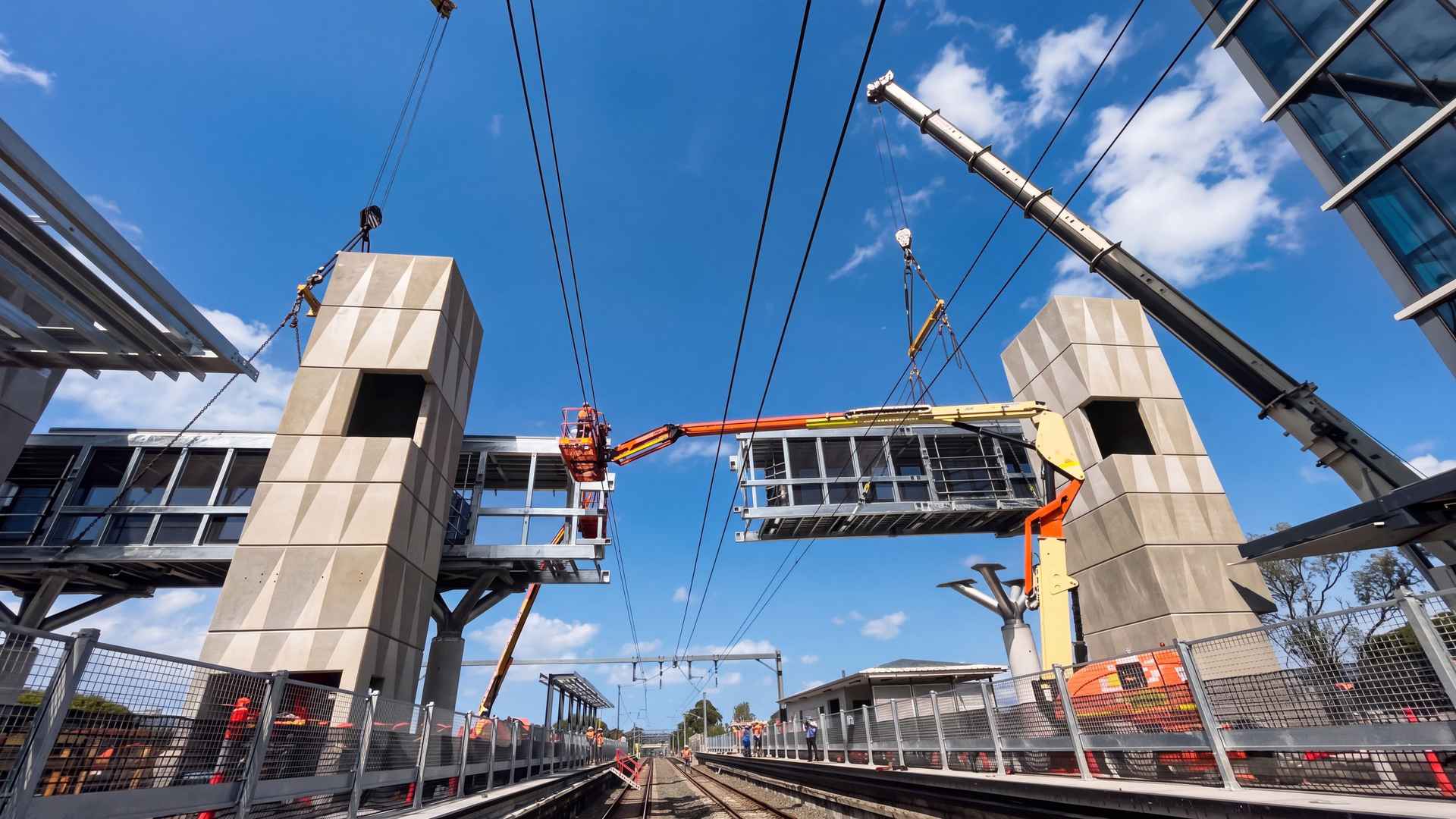 Two mobile cranes lift a new steel footbridge span between concrete lift towers over live railway tracks